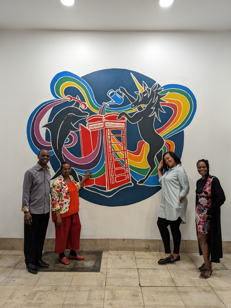 Four people from Hillview Community Services stand by mural of a unicorn and a dolphin watering a phonebox with rainbow swirls