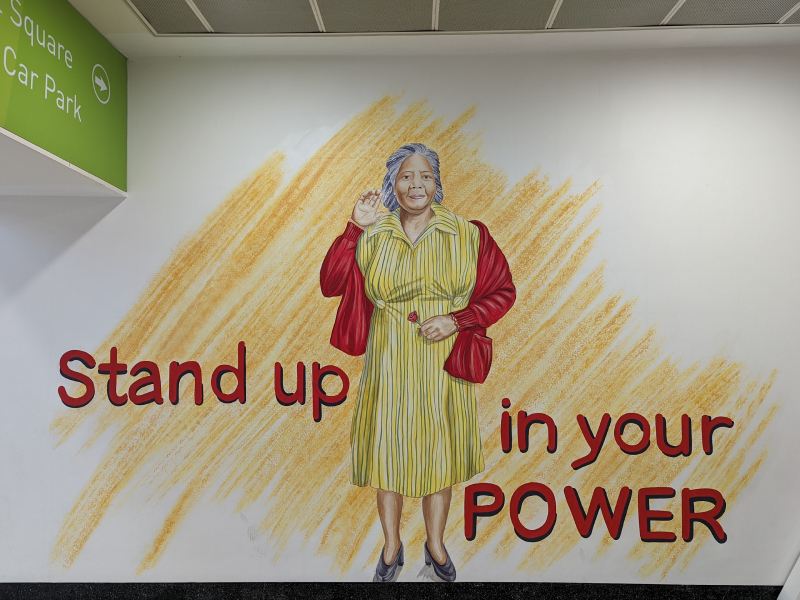 Painting in the likeness of Sybil Phoenix, older woman wearing a yellow dress. Stand up in your Power is written around her.