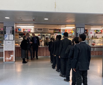 queue of school boys in a lunch hall with three adults standing to the left of them talking
