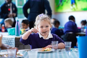 Young child in purple school uniform looking at camera and eating their school lunch happily