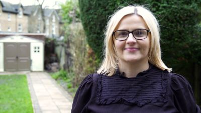 A white woman wearing a black blouse, wearing black-rimmed glasses, with blonde shoulder length hair