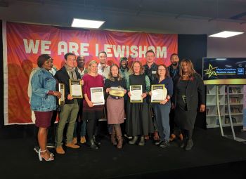 A mixed group of men and women - different ages and ethnic backgrounds - standing on a stage holding framed certificate with orange flag behind them which says We Are Lewisham