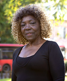 A black women with curly black and brown hair stands against a blurred out background which includes a red single decker bus and leafy green trees