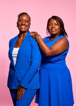 Two middle-aged Black women wearing bright blue. One is in a trouser suit, the other is in a dress
