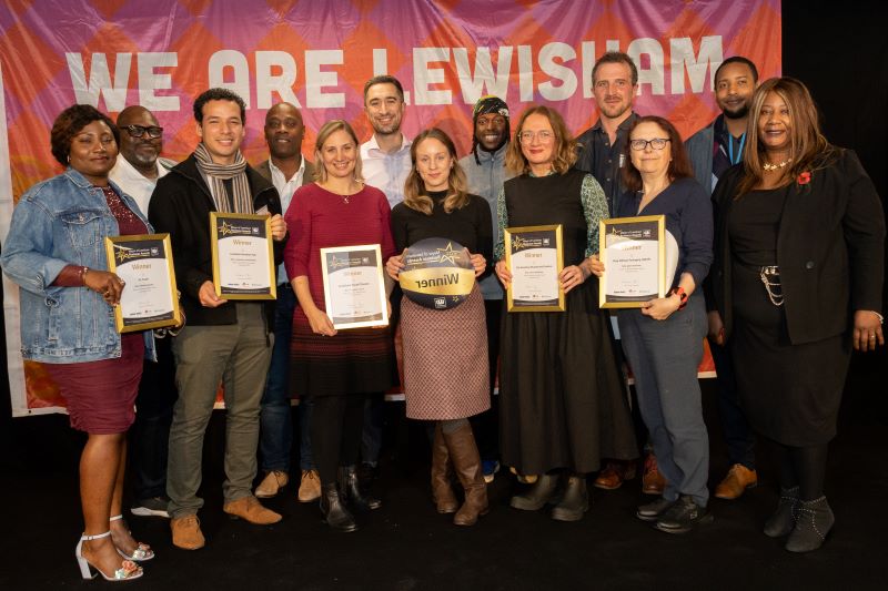 A group of people standing on a stage - they a re a mix of ages, genders and ethnic backgrounds and are all holding framed winners certificates.