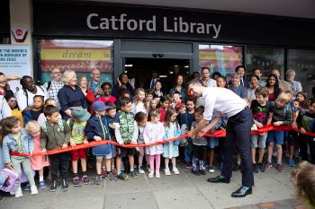 The Mayor - a man in his 30s - wearing a white shirt and black trousers cuts a ribbon which is being held by lots of small children. There are two rows of adults behind the children. And behind them is Catford Library - the sign is white text on a black background.