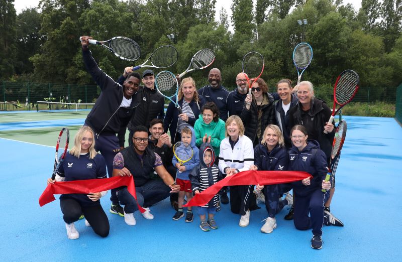 A group of around 20 people standing on a blue tennis court. They are a mixture of ages, genders and ethnic backgrounds. They are all holding tennis rackets in the air. The people who are in the front row are kneeling and appear to have just cut a red ribbon. 