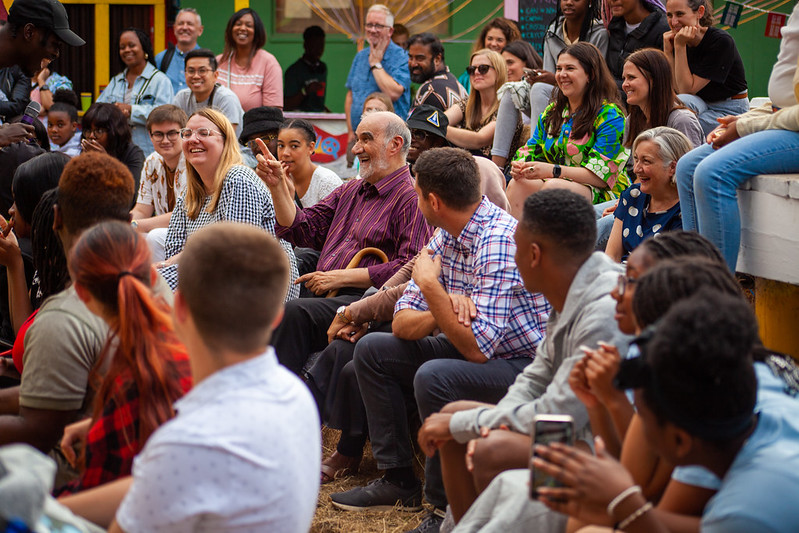 Lots of people - different ages, genders and ethnic backgrounds - sitting down. They are in the audience for an outdoor event. There is straw on the floor. Everyone is laughing. A young black man holds a mic to the mouth of an older white man