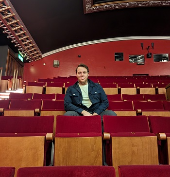 White man with short brown hair, wearing blue denim jacket seated among rows of red theatre seats in a theatre auditorium