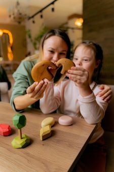 A woman sits smiling at a table with a little girl who has Down's Syndrome. The little girl is breaking a gingerbread biscuit which is being held up to the camera