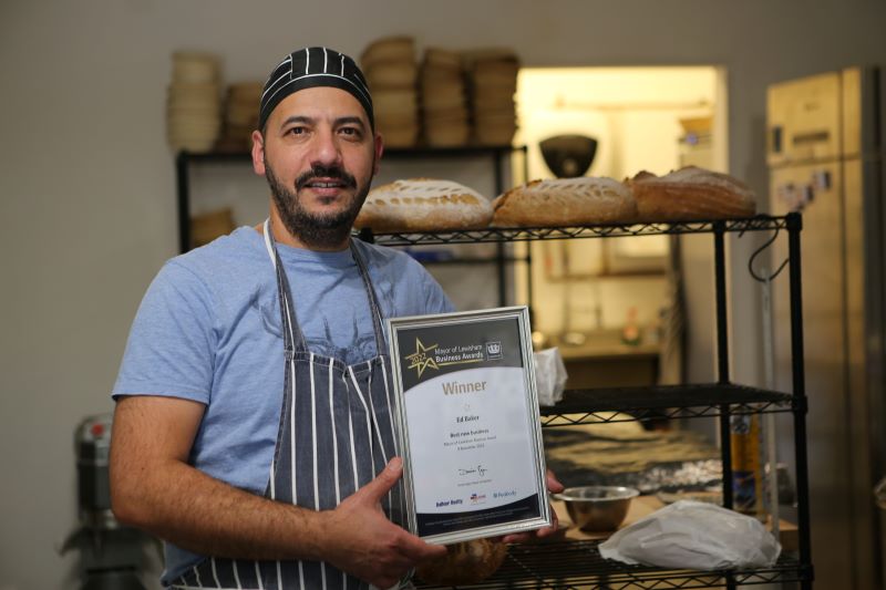Image of man in matching blue and white striped apron with shelves of bread behind him, holding a framed winners certificate
