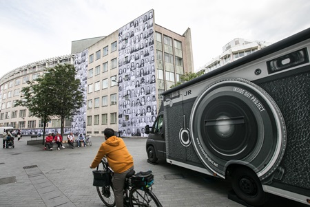 A cyclist cycles past a lorry that looks like a large digitial camera. Inside Out's portraits are displayed on a building in the background. 