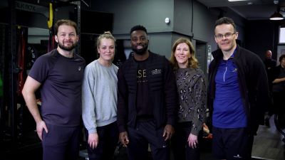 Five people standing in a gym. They are a mix of genders but all are white except the person in the middle who is a young black man, wearing a black outfit - his top sayd 'Base 13' in gold letters
