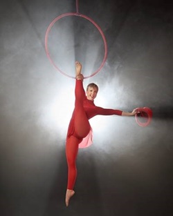 A lady in red hangs from a trapeze holding  her ankle