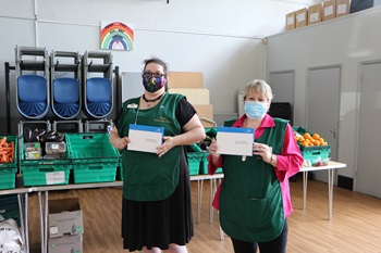 Two Evelyn Community Store volunteers holding a rapid test kit looking at the camera. They are wearing masks and green aprons.
