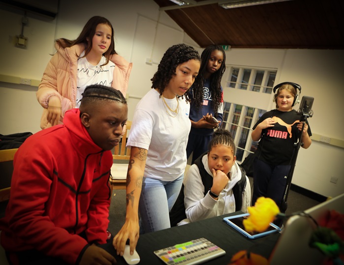 A group of young people standing around a computer. One of them is wearing recording headphones and standing by a studio microphone.