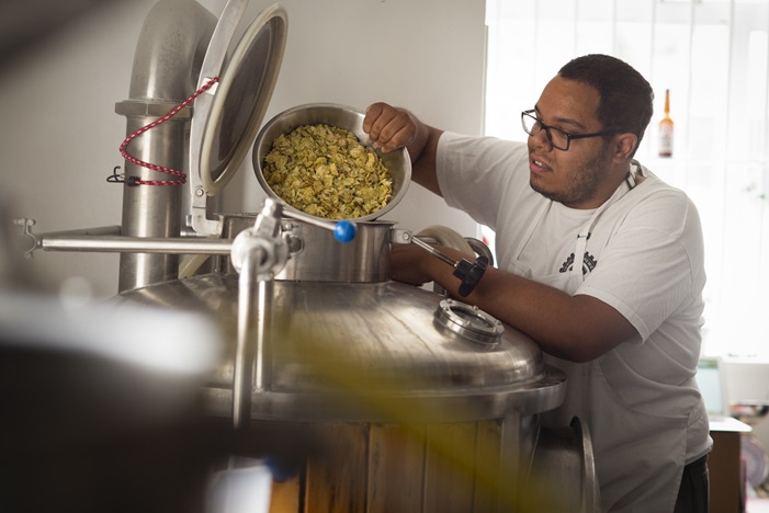Person pouring hops into a beer fermenter. They are wearing a white shirt and white apron.