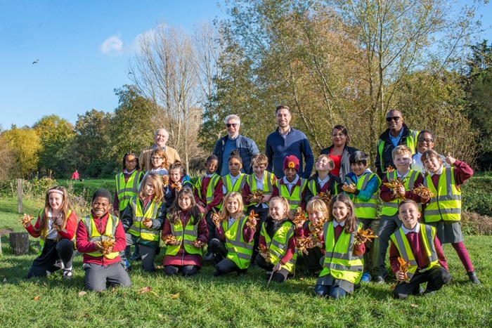 A group of 20 school children in a park, with five adults standing behind them. The children are wearing high-vis jackets and holding bunches of leaves. The weather is sunny.