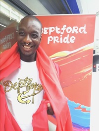 Ste Rikhardsson wearing a Deptford t-shirt standing in front of a Deptford Pride poster. 