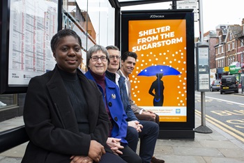 Left to right: Dr Catherine Mbema, Director of Public Health Lewisham Council; Cllr Chris Best, Deputy Mayor and Cabinet Member for Health and Adult Social Care; Andrew Jordan and Pierre Rogier from JC Decaux. All sat at a bus stop infront of a Lewisham Sugar Smart bus stop advert.