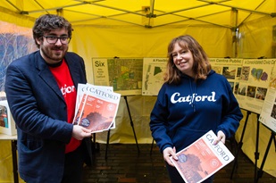 Two people in Catford jumpers holding magazines