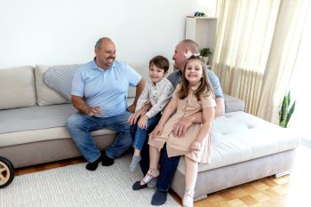 A couple - two bald white men with beards and moustaches - sitting on a sofa with two young children. One is a girl wearing a beige dress, the other is a boy wearing a white shirt and jeans