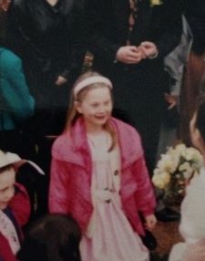 Image of a young girl wearing a pink jacket and dress. She is surrounded by other people who are all partially obscured