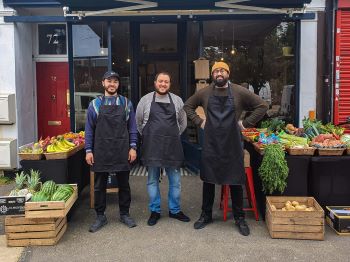 The three founders of Marvellous Greens and Beans standing outside their shop on Brockley Rise, surrounded by crates of fresh fruit and veg