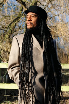 Maxi Priest is standing in front a low wood and wire fence. There is a tree beyond the fence