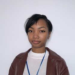 A young, Black woman standing in front of a white wall wearing a brown leather jacket and white t-shirt underneath.