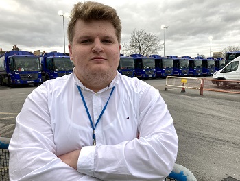 A young man in a white shirt with his arms crossed standing in front of refuse trucks
