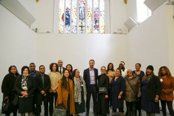 A large group of people - different ages and genders - from a variety of different ethnic backgrounds. They are standing in front of a white wall which has a large stain-glass window