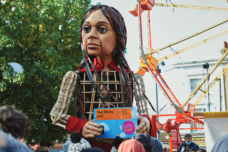 12 ft puppet of Syrian girl, Little Amal walking through Lewisham, carrying a Lewisham Library card