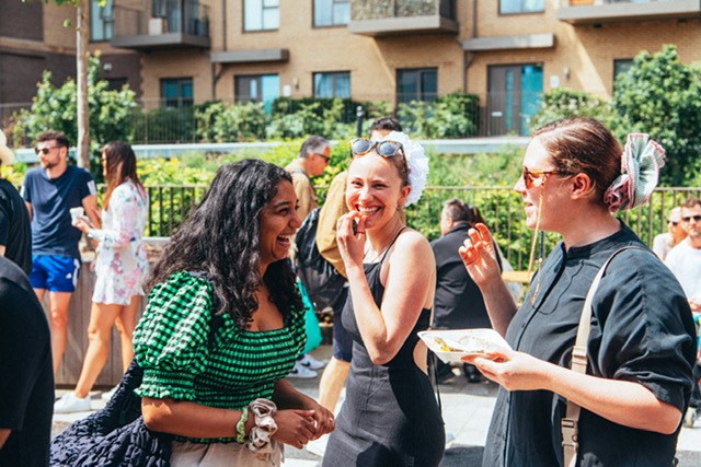 Three young women - two are white with brown hair, they are both wearing black tops, one is brown-skinned, possibly of South Asian origin, with long black wavy hair and is wearing a green top with black stripes. They are laughing while eating.