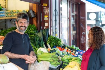 A male business owner stands between the fruit and vegetable tables at the front of his shop. He is talking to Cllr Powell - a woman with long brown hair 