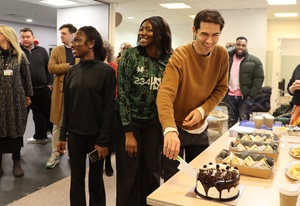 A man cuts a cake and smiles while a group of people stand near him