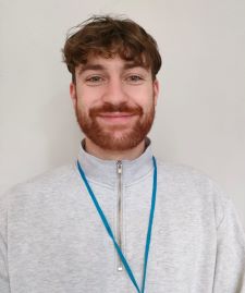 A smiling, young man with beard, wearing white shirt standing in front of a white wall