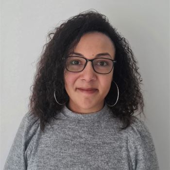 Young woman with shoulder length brown curly hair, with silver hoop earrings, wearing a grey roll neck sweater, standing in front of a white wall