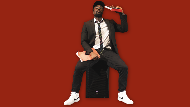 A young, Black man wearing a suit is sitting on a speaker. He is holding a text book in one hand and a keyboard in the other