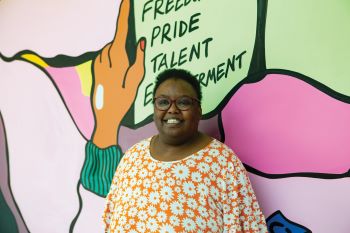 Black woman, with short black afro hair, wearing a yellow top stands in front of a mural which has lots of bright colours