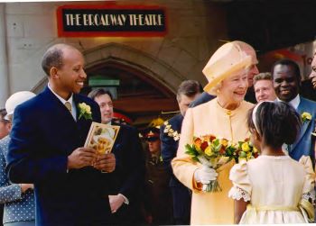 Her Majesty The Queen meets crowds including a young girl, who has her back to camera, outside of a building which has the words The Broadway Theatre above the door.  The Queen is  wearing matching lemon hat and longline jacket. The picture was taken when the Queen visited in Catford in 2002 for the Golden Jubilee.