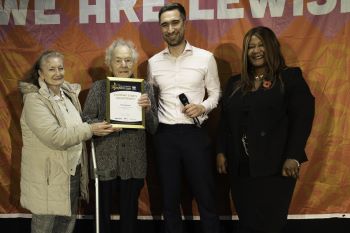 Two men and two women standing on a stage - the older of the two men (who is elderly) is holding a framed certificate - with orange flag behind them which says We Are Lewisham