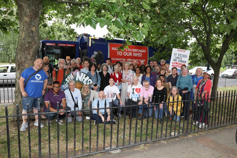 A group of people stand in a park with save Lewisham Hospital banners in the background