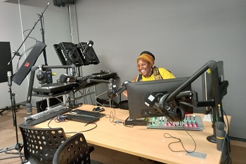 A Black woman with a yellow scarf and dreadlocks piled up on her head sits behind mixing desk with mixer, laptops, microphones and other sound equipment.