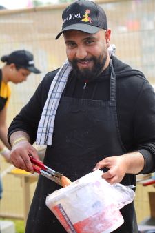 A man with a black beard, wearing a black cap and black apron, mixing something in a large container is looking at the camera