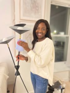A Black woman with long, black straight hair. She is wearing a long-sleeved white top and blue jeans and is standing in front of two large framed pictures on a white wall. She is using a duster on two uplight lamps