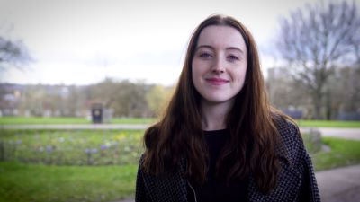 A young white woman with long dark brown hair, wearing a dark coat and top. She is standing in a park.