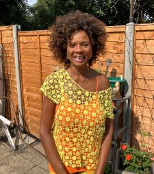 A black woman with brown curly hair and a yellow, off-the-shoulder top stands smiling in front of a wooden garden fence.